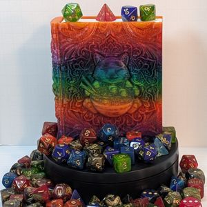 A dice box shaped like a tome. It's surround by colorful TTRPG dice. The box is printed out of a shiny rainbow gradient filament, and it has a happy cat sitting behind a bowl of ramen.