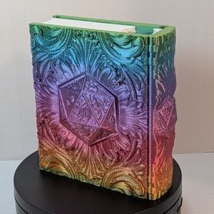 A 3D printed "tome", printed out of a shiny rainbow silk filament. On the front is a d20, showing the 20. There is a white insert for the "pages" and it is turned so you can see the matching designs on the back and the spine.