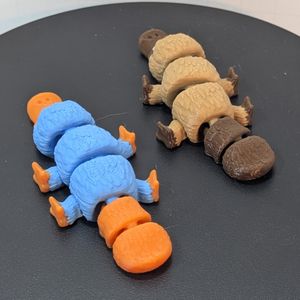 A light brown platypus with darker brown bill and feet, next to a blue platypus with orange bill and feet. They are photographed from the back, showing the articulation in their tails.