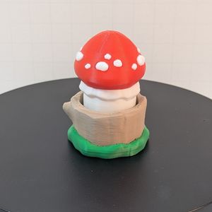 A 3D printed mushroom with a red cap. There are white spots around the red cap, and the stem is white, as well. The base looks like a light tan tree stump, with green around the bottom. 