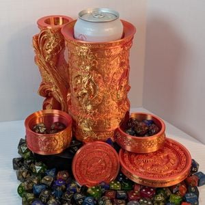 A reddish-gold 3D printed tankard, with an ornate handle. The artwork on the side is of a dragon head, and there's a tray sticking out the bottom front. It's disassembled, with the dice vault beside it, a can in the tankard, and dice everywhere.