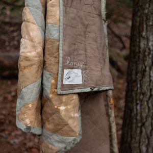 The Honey Bee Quilt hanging in the crook of a young tree, showing the flame & honeycomb label and the name of the quilt hand embroidered in undyed organic cotton thread