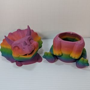 A chunky little baby triceratops, 3D printed out of a bright rainbow PLA plastic. The head and shoulders are sitting beside the body, which shows the opening for the dice vault and set of magnets that keep it in place.