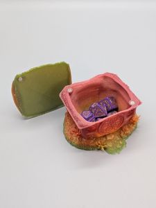 A 3D printed dice vault in the shape of the home of a halfling. It's got grass on the roof, a round chimney, and a round door. The top is off, showing the magnets and how the dice fit in.