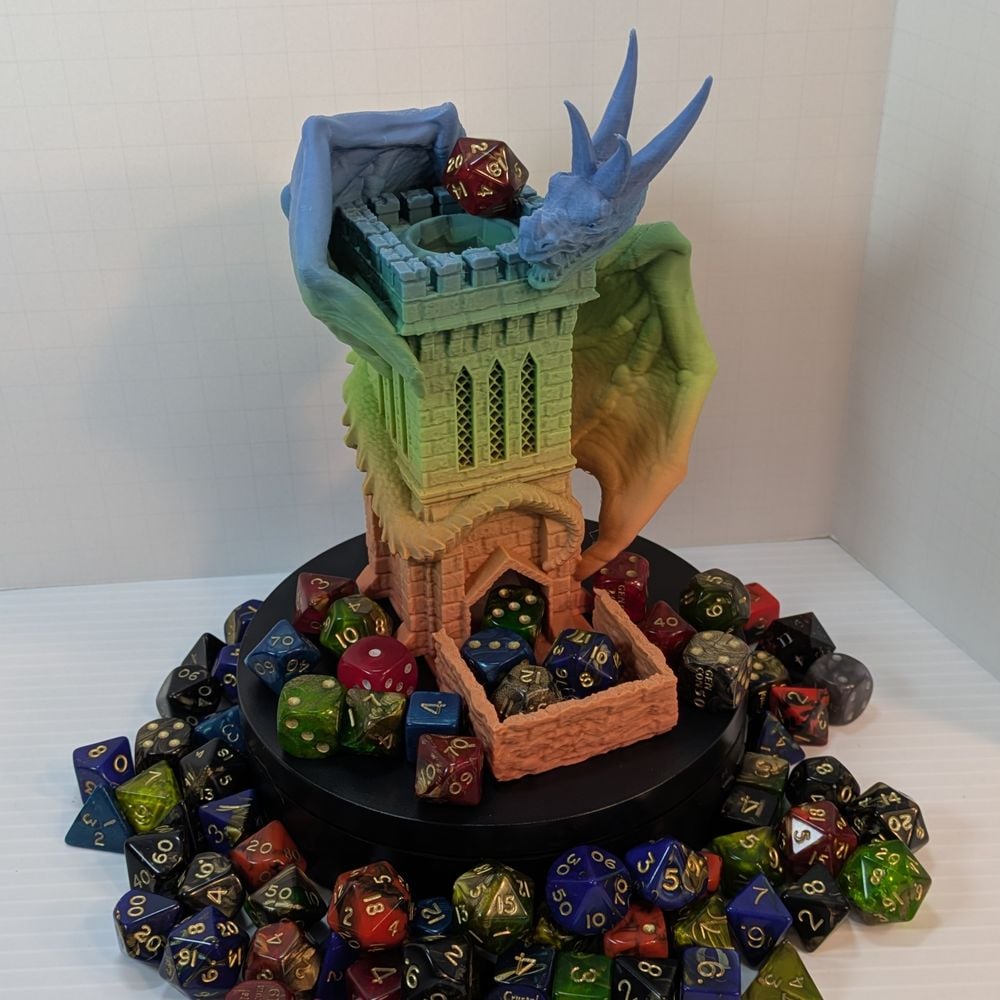 A 3D printed dice tower in the shape of a square stone tower with rock walls as a catching tray. Wrapped around the tower is a wyvern with four sharp horns. It's in a matte rainbow filament. It is surrounded by dice.