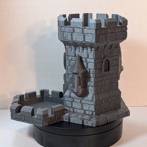 The side of the 3d printed dice tower, shaped like a castle tower. It's a metallic steel color, and it's shaped like uneven stonework with a tray at the bottom that matches the top.
