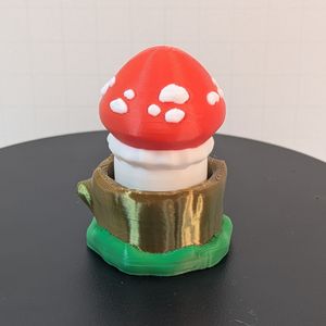 A 3D printed mushroom with a red cap. There are white spots around the red cap, and the stem is white, as well. The base looks like a shiny brown tree stump, with green around the bottom. 