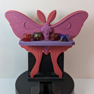 A small 3D printed shelf in the shape of a Luna Moth. It is a pinkish-purplish hue, and the shelf across the front has some standard size TTRPG dice on it.