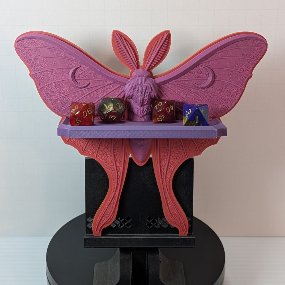 A small 3D printed shelf in the shape of a Luna Moth. It is a pinkish-purplish hue, and the shelf across the front has some standard size TTRPG dice on it.