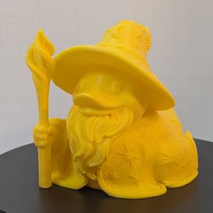 A yellow 3D printed rubber duck inspired piece dressed like a fantasy wizard, with a cloak and hat covered in stars and moons, a twisted wood staff, and a long beard under the bill.