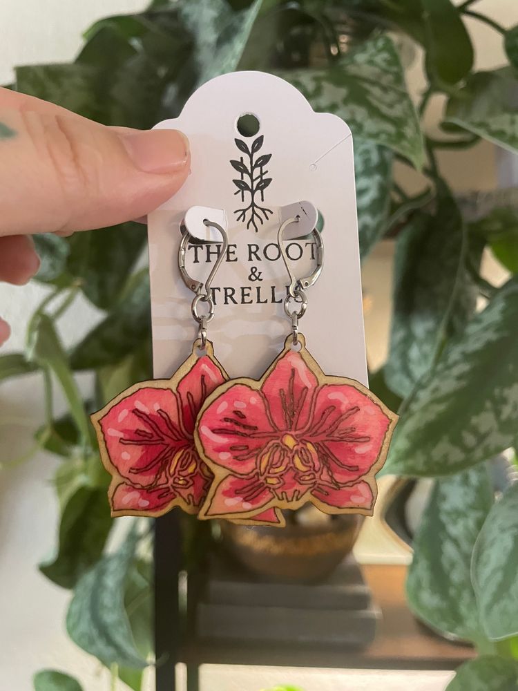 An image of a pair of pink earrings with an orchid design being held by the hand of a person in front of a botanical backdrop.