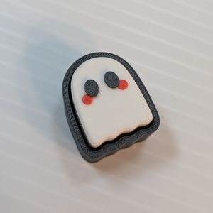 The 3D printed Ghost clicker, with no chain. It's a white ghost, with black eyes and red cheeks. It's inside a black case that surrounds it and hides the keyboard switch, and gives the ghost a black outline.