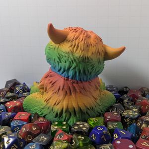 The back view of a chubby little 3D printed owlbear, printed in a rainbow gradient PLA. The little guy is surrounded by colorful TTRPG dice.