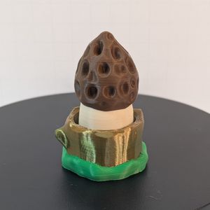 A 3D printed morel mushroom. The top is brown, on a light tan stem. The tree trunk--styled base is a shiny silk earth tone, with green ringing the bottom to look like grass.