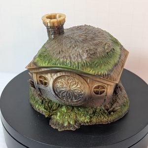 A 3D printed dice vault in the shape of the home of a halfling. It's got grass on the roof, a round chimney, and a round door. The top comes off to put the dice in. This one is a lighter green gradient.