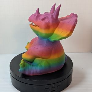 A chunky little baby triceratops, 3D printed out of a bright rainbow PLA plastic. There is a seam at the shoulders where you can lift the head off and use it as a dice vault.