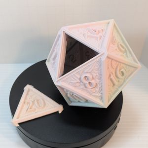 The 3d printed d20 dice vault, resting on its own on a black base. Once again, the 20 is removed to show the triangular opening for the dice to go into, and the color is going from a pastel pink to a light blue.