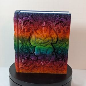A 3D printed tome, with a chubby baby triceratops, Chomper, on the cover. Comper is surrounded by decorative scrollwork. The tome is printed out of a shiny rainbow gradient filament.