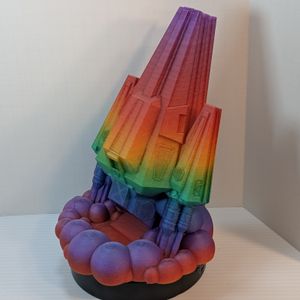 A 3D printed dice tower in the shape of a space ship. The main body is a wedge, and the thrusters on either side are wedges. It's in a rainbow gradient filament, and the dice come out in the cloud of smoke under the thrusters.