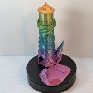 A 3D printed dice tower in the shape of a lighthouse, with a rocky base . It's printed in a very shiny rainbow filament. The dice catcher tray is sitting off to the side, showing how it slides out of the tower.