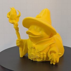 A yellow 3D printed rubber duck inspired piece dressed like a fantasy sorcerer, with a pointed hat, a staff with a crystal on the end, vials on their belt, and a small ball of flame in their hand.