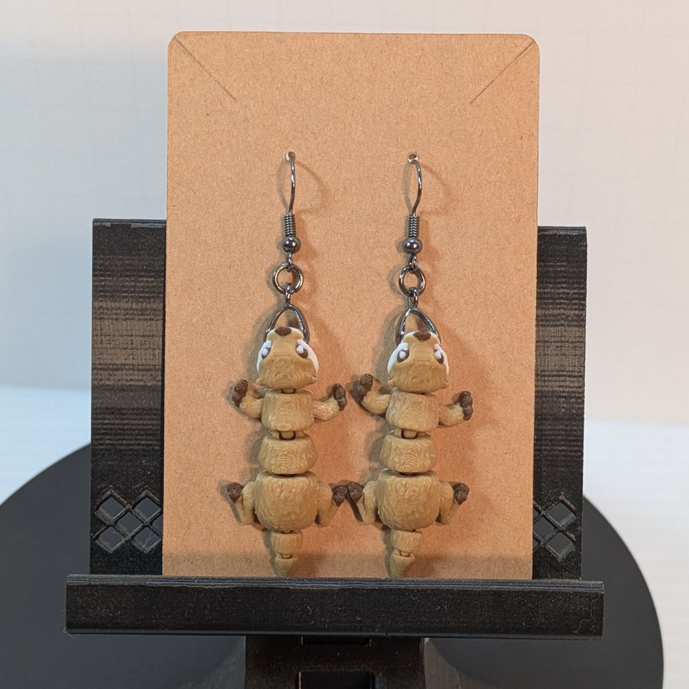 A pair of 3D printed earrings in the shape of River Otters. They're tan, with white and brown markings. They're on fish hook-style earring hooks and hanging on their cardboard card.