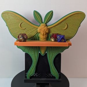 A small 3D printed shelf in the shape of a Luna Moth. It is a greenish-yellowish hue, and the shelf across the front has some standard size TTRPG dice on it.