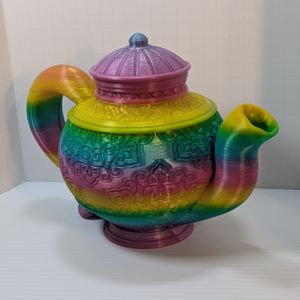 A 3D printed teapot, printed out of a rainbow gradient filament. It has ornate markings "carved" into the sides, with a cap on top. The handle flares out the back, and the spout stays close to the body.