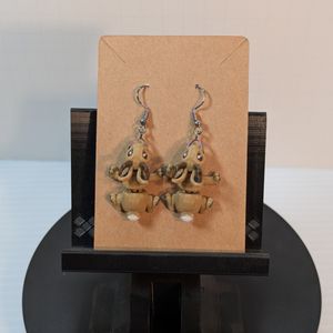 A pair of 3D printed earrings, shaped like Jackalopes. They are light brown, with dark brown prongs, and they're attached to rainbow colored stainless steel earring hooks. The are attached to the cardboard card they will ship on.