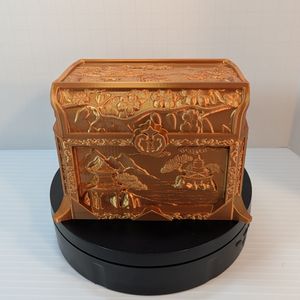 A copper colored 3D printed dice box, in the shape of a chest. There is a fake lock on the front, with images of Chinese architecture and trees on water.