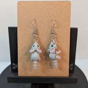 A pair of 3D printed earrings in the shape of a gray mouse, hanging from silver fishhook earrings on a brown cardboard earring card.
