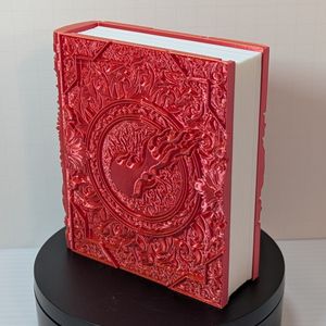A 3D printed box that's shaped like a tome with fancy scrollwork and a ball of flame on the cover. It's made out of a shiny red/yellow filament, and is turned so you can see the white "pages" insert in the center, with uneven pages between the covers.