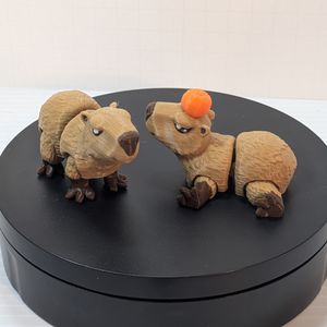 A pair of smaller 3D printed Capybaras, printed in a tan filament for the coat and a darker brown for the nose and feet. One is standing, the other laying down with a Yuzu fruit on its head.