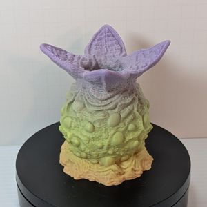 A 3D printed dice container that looks like an alien egg. It's lumpy, and there are four wedges open at the top, where you can drop in your dice. This starts with an off-yellow on the bottom, going up to green, then purple at the top.