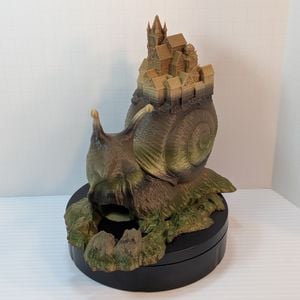 A slight front angle of a 3d printed dice tower in the shape of a snail with a town on its shell. The dice exit into a tray that looks like rocky ground from the front of the snail.
