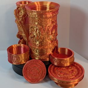 A reddish-gold 3D printed tankard, with an ornate handle. The artwork on the side is of a dragon head, and there's a tray sticking out the bottom front to catch dice. The dice box is beside it, and all lids are removed.