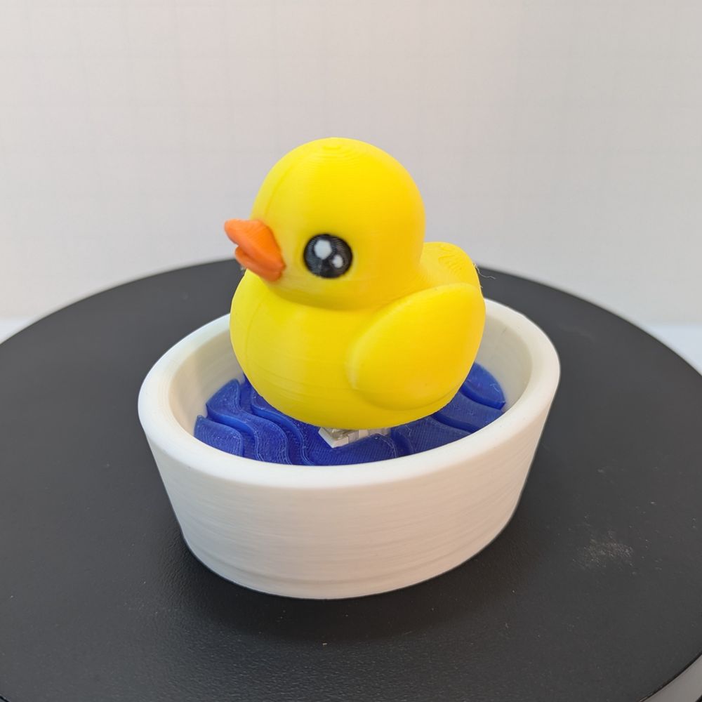 A 3D printed yellow ducky with an orange bill, on top of a small white bowl filled with blue "water." You can just barely see the mechanical keyboard switch between the duck and the water.