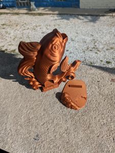 A 3D printed dice tower in the shape of a koi. It's in a bronze filament, and the fish looks like it's resting on it's tail. The head is up, with the mouth open, which is where you drop the dice. The dice exit from the base of the tail.