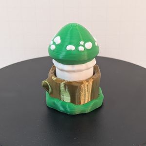 A 3D printed mushroom with a green cap. There are white spots around the red cap, and the stem is white, as well. The base looks like a shiny light brown tree stump, with green around the bottom. 