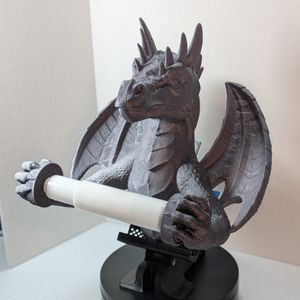 A dark metallic-silver colored 3D printed toilet paper holder in the shape of a dragon. The TP is held between its outstretched arms, with outstretched wings in the back.