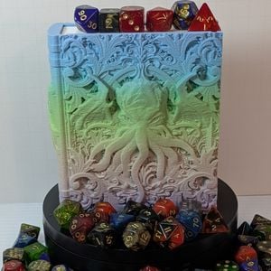 A dice box in the shape of a tome. It's printed in a muted gradient that goes from light blue, green, pink, to purple. On the cover is a face with many tentacles coming out of it. It's surrounded by dice while resting on a black stand.
