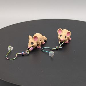 A pair of 3D printed mouse earrings. They are printed in color, with pink on their ears, toes, and nose, black and white eyes, and a light brown fur. They look like they are smiling, with the earrings in their mouths.