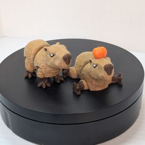 A pair of smaller 3D printed Capybaras, printed in a tan filament for the coat and a darker brown for the nose and feet. One is standing, the other laying down with a Yuzu fruit on its head.
