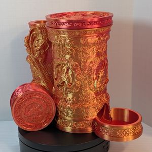 A reddish-gold 3D printed tankard, with an ornate handle. The artwork on the side is of a dragon head, and there's a tray sticking out the bottom front to catch dice. Leaning against the side is a circular dice box with the same art on the lid.