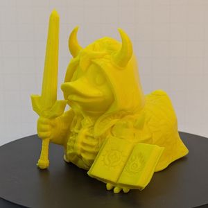 A 3D-printed duck in the style of a rubber duck. It's dressed as a warlock, with an open tome in one hand, and a large sword in the other. The breastplate looks like a rib cage, and they're wearing a horned hood.