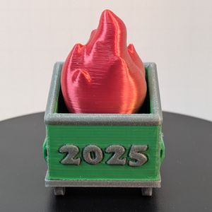 A 3D printed dumpster fire. The dumpster is green with a silver base and rim around the top, and the fire coming out the top is a shiny red with yellow accents. Across the front of the dumpster is "2025" in the same silver as the base and rim.