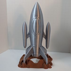 a 3D printed dice tower in the shape of a silver retro-style rocket ship. It's shaped like a pointed cylinder, with two smaller pointed cylinders on the side. On the bottom around the rocket exhaust are four fins that rest in holes on the rock-like base.