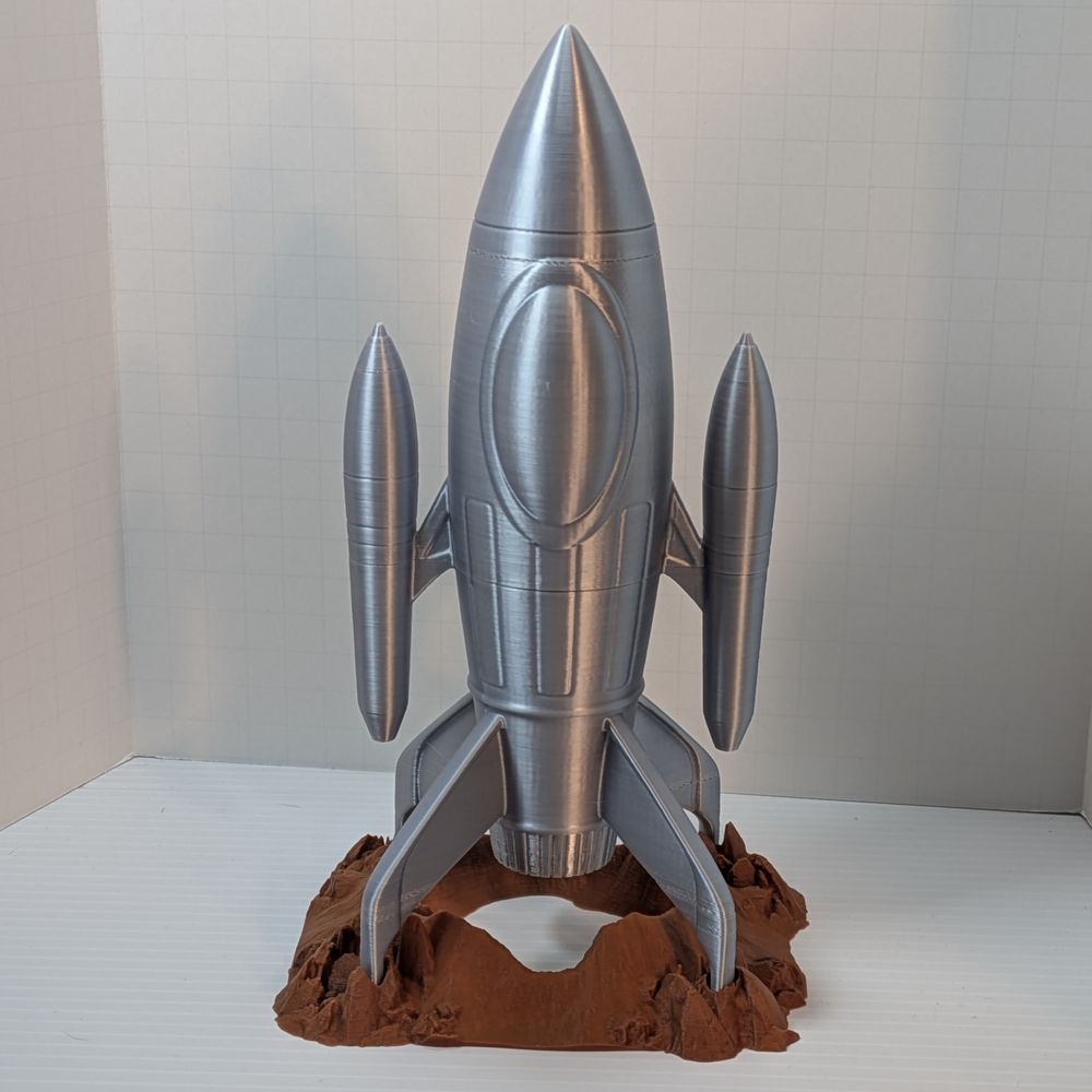 a 3D printed dice tower in the shape of a silver retro-style rocket ship. It's shaped like a pointed cylinder, with two smaller pointed cylinders on the side. On the bottom around the rocket exhaust are four fins that rest in holes on the rock-like base.