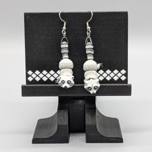 3D printed earrings in the shape of raccoons, printed in color. They are hanging from silver "fish hook" earrings attached to their tails.