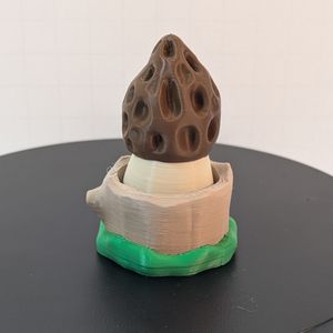 A 3D printed morel mushroom. The top is brown, on a light tan stem. The tree trunk--styled base is a different shade of tan, with green ringing the bottom to look like grass.